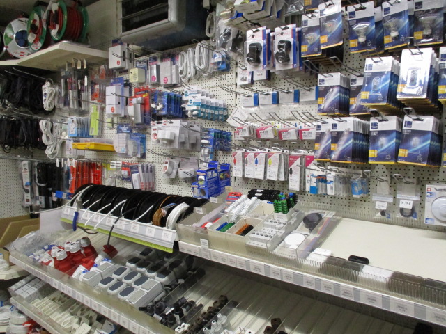 Stecktafelwand mit verschiedenen Elektroartikeln wie Kabeln, Adaptern, Batterien, Steckern, Schaltern und verpackter Elektronik, die in einem Baumarkt in Regalen und an Haken aufgehängt sind.