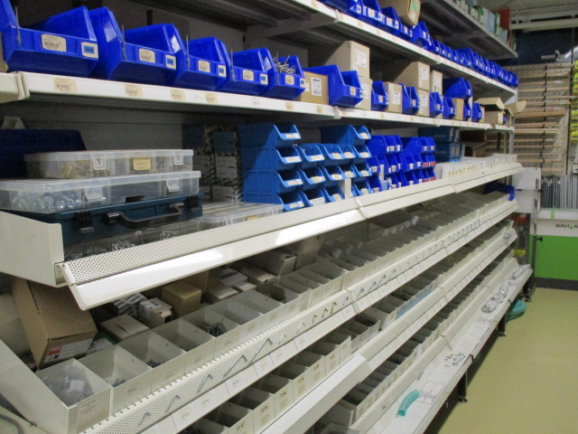 Ein Ladengang mit Regalen mit blauen Plastikbehältern, von denen einige beschriftet und mit kleinen Eisenwaren gefüllt sind, während viele Behälter im unteren Regal meist leer sind. Der Boden ist hell, und die Regale sind aus Metall.