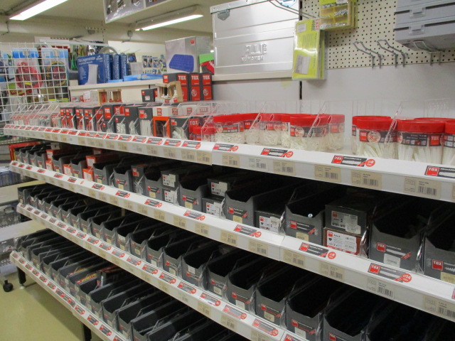 In einem Ladenregal sind verschiedene Eisenwaren in Plastikbehältern und -kisten nach Typen geordnet, wobei einige Behälter rote und weiße Container enthalten. Die Regale sind beschriftet, und im Hintergrund sind weitere Produkte zu sehen.