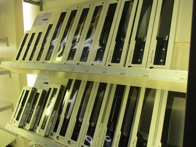 Zwei Regale mit verschiedenen Küchenmessern in weißen Verpackungen, unter denen jeweils Etiketten und Preise sichtbar sind, vor einer hell erleuchteten Auslage.