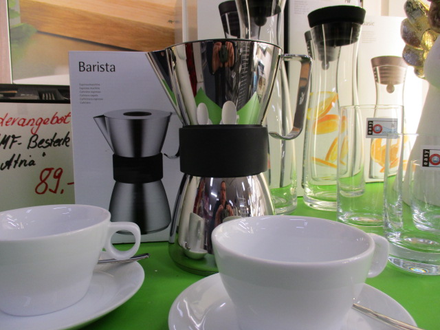 Eine glänzende Kaffeemaschine aus Metall mit der Aufschrift Barista wird mit ihrer Schachtel zwischen zwei weißen Tassen und Untertassen, zwei klaren Gläsern und anderen Glaswaren auf einer grünen Fläche ausgestellt. Die Preisschilder sind teilweise sichtbar.