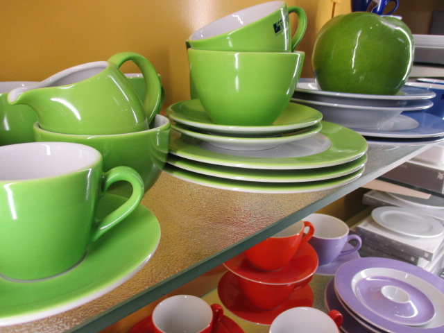 Auf einem Regal stehen grüne Teetassen mit Untertassen und Tellern sowie eine grüne Teekanne und ein großes grünes Apfelornament. Darunter sind rote, orangefarbene und violette Teetassen und Untertassen fein säuberlich angeordnet.