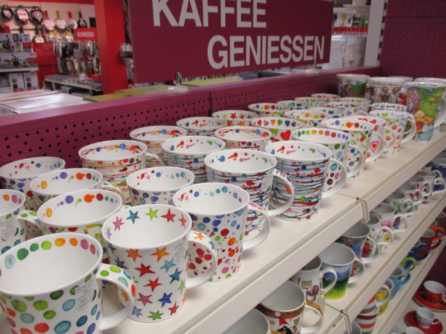 In den Regalen stehen bunte Tassen mit verschiedenen Mustern, darunter Tupfen, Streifen und Sterne. Darüber steht ein Schild mit der Aufschrift KAFFEE GENIESSEN, und im Hintergrund sind weitere Küchenartikel in einem hell erleuchteten Laden zu sehen.