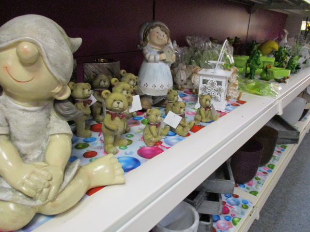 Ein Ladenregal mit Keramikfiguren, darunter ein lächelndes Kind und mehrere Teddybären, neben anderen Dekorationsartikeln und kleinen Topfpflanzen auf einer bunt gepunkteten Oberfläche.