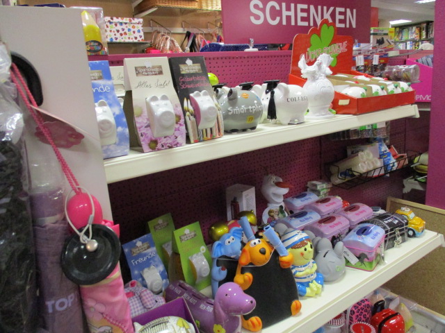 In den Regalen eines Geschäfts sind verschiedene bunte Sparschweine, Geschenkartikel und Spielzeuge ausgestellt. Auf einem rosa Schild über den Regalen steht SCHENKEN. Die gesamte Umgebung wirkt fröhlich und gut organisiert.