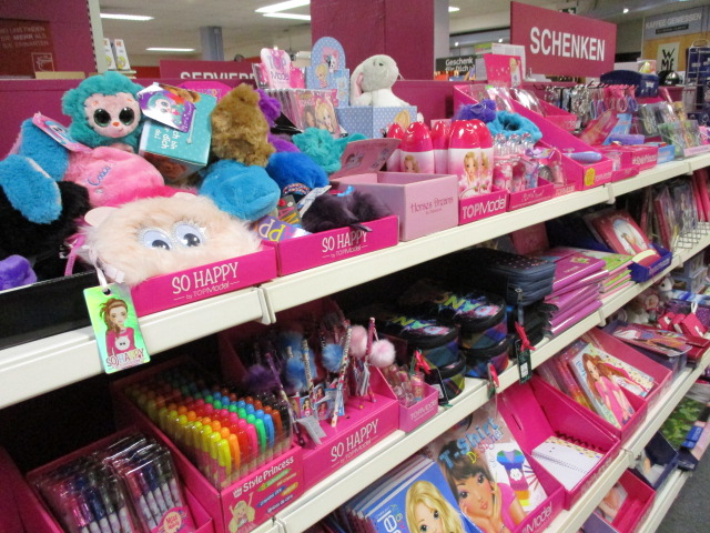 Ladenregal mit buntem Spielzeug, Plüschtieren, Stiften, Notizbüchern, Mäppchen und verschiedenen Schreibwaren, meist in Rosa und mit der Marke Barbie, die sich an Kinder richten. Über dem Regal sind Schilder in deutscher Sprache zu sehen.