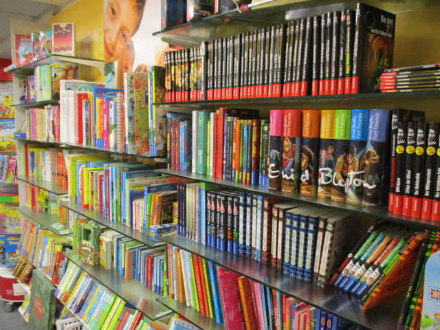 Ein hell erleuchtetes Buchladenregal, gefüllt mit bunten Kinderbüchern, Graphic Novels und Serien, darunter auch sichtbare Enid Blyton-Titel, ordentlich auf Metallregalen angeordnet.