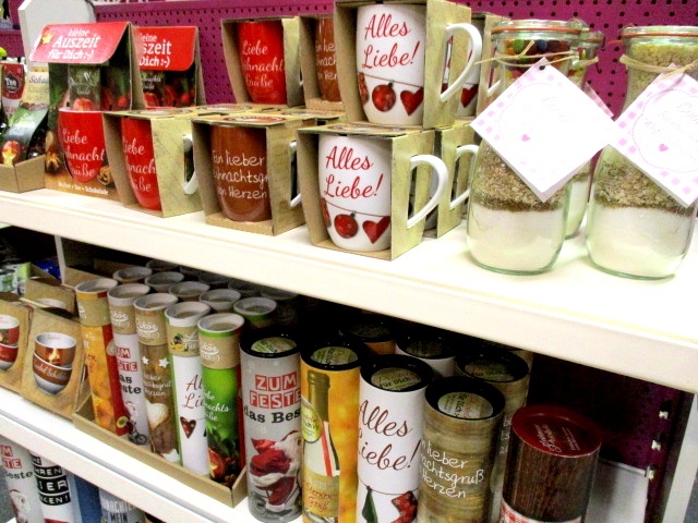 In einem Regal im Kaufhaus Heitmüller stehen bunte Tassen mit deutscher Aufschrift, Gläser mit geschichteten Backzutaten und verschiedene verpackte Lebensmittel, alle fein säuberlich in Reihen angeordnet.