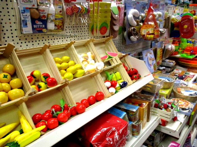 Ein Regal im Kaufhaus Heitmüller zeigt buntes Spielzeugobst und -gemüse in hölzernen Behältern, während im Hintergrund verschiedene Spielzeuglebensmittel und Küchenaccessoires von Haus&Co an einer Stecktafel hängen.