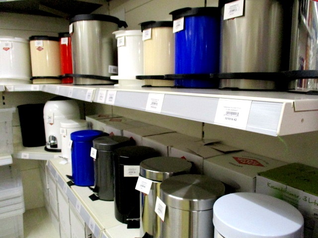 In einem Regal im Kaufhaus Heitmüller stehen verschiedene Mülleimer aus Metall und Kunststoff in unterschiedlichen Farben, Größen und Formen. In diesem gut sortierten Fachgeschäft sind die Preisschilder im Regal sichtbar.